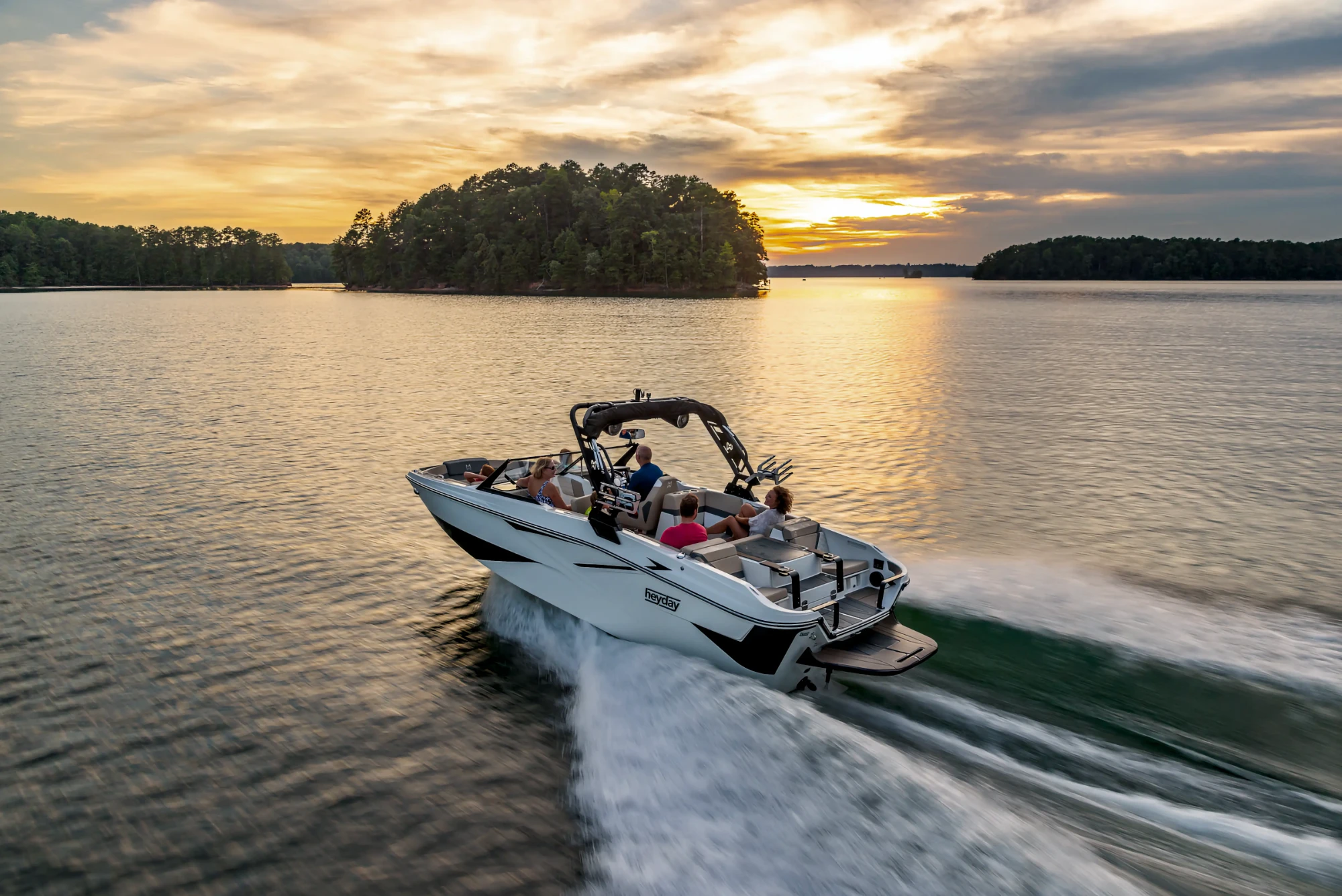 A white Heyday wake boat with passengers cruising across a lake at sunset